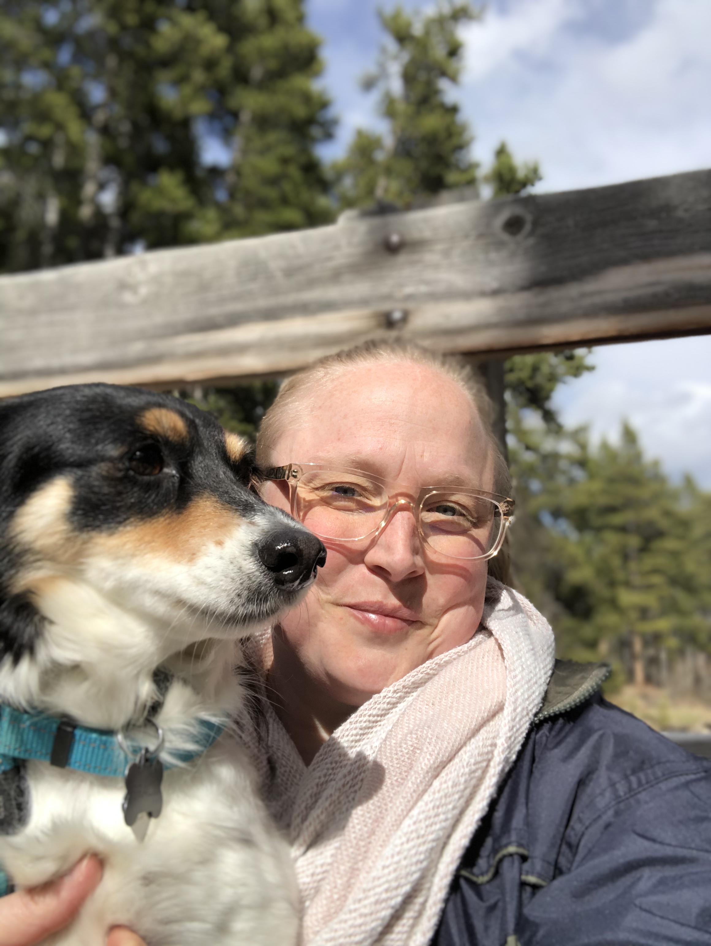 Teo sits on a bridge in Montana with trees behind them. Teo holds Gracie, a rescue pup. Teo smiles wearing clear glasses and a big scarf.