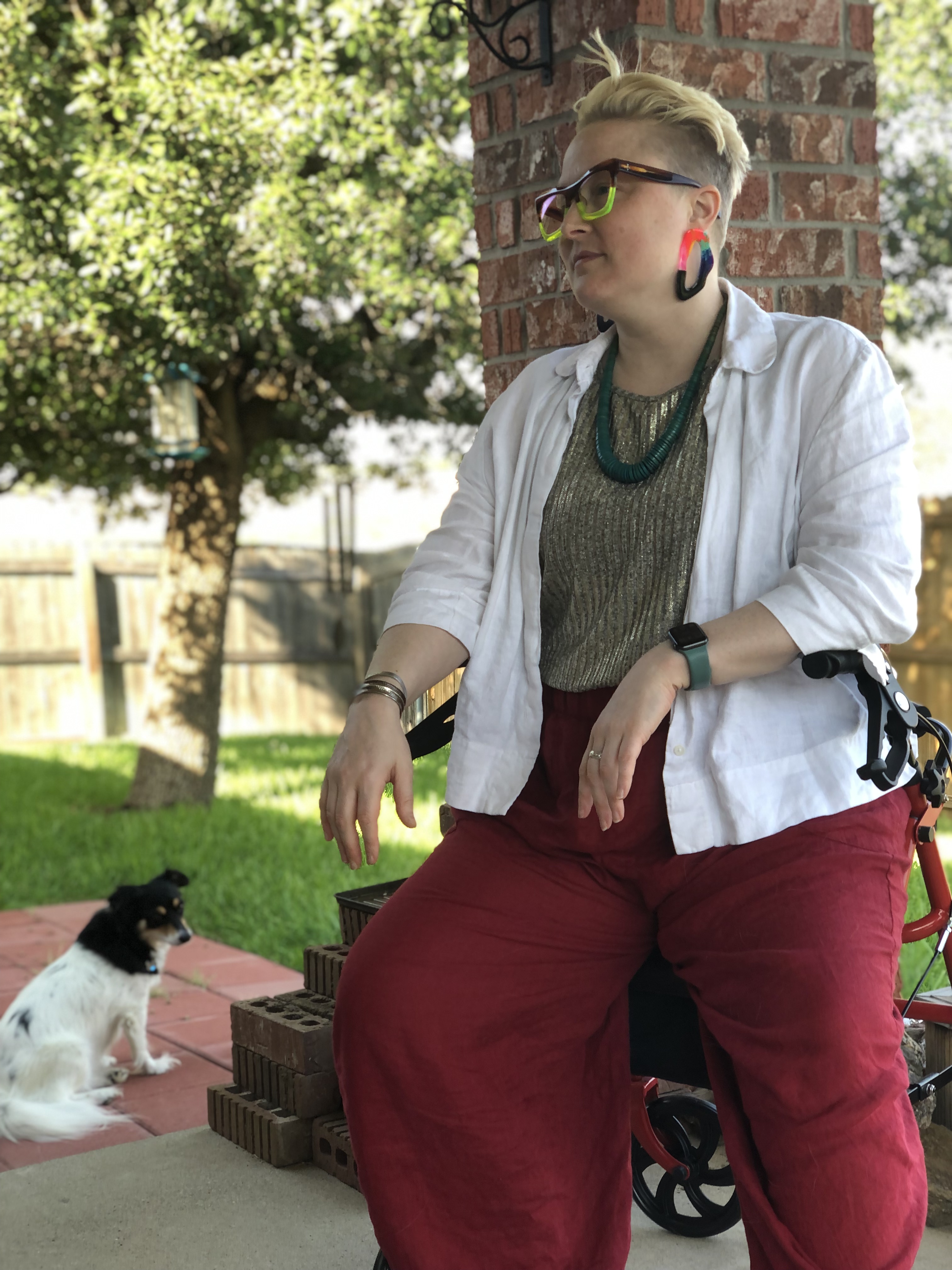 Teo sits outdoors with pup Gracie behind them. Teo is wearing a stylish outfit of red linen pants, a gold lame tank, and white linen blazer. Teo is styling with bold blue glasses, a teal wood necklace, and large bright pink and black acrylic earrings.