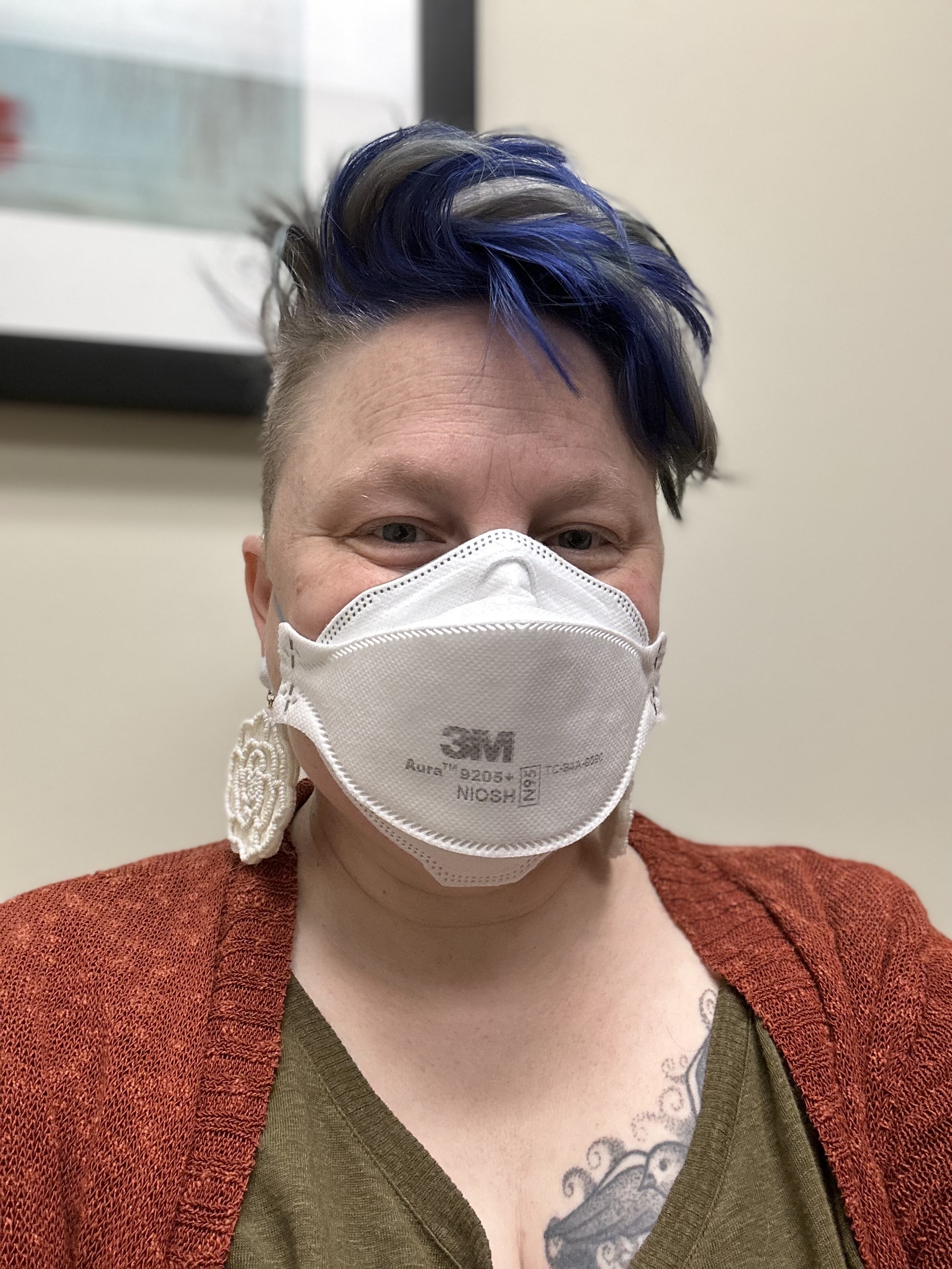 Teo wears a mask, large white earrings, a red cardigan, and green tshirt. Teo's hair is a bright purple and their tattoo is slightly visible on their chest.