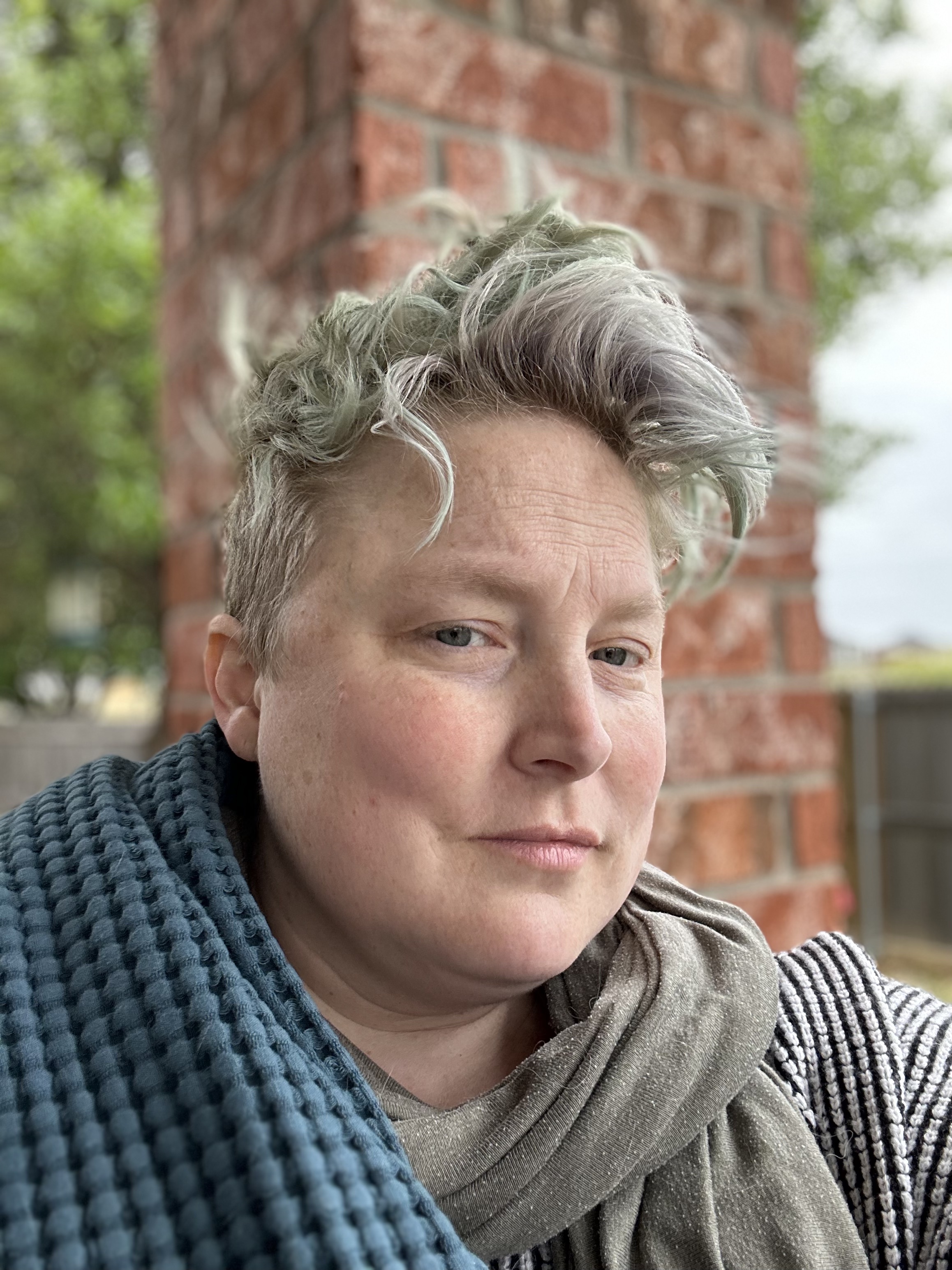 Teo sits outside wearing a grey/green scarf and blue waffle blanket. Teo's hair has faded from a dye job to a blue/green color and is sitting wildly on their head