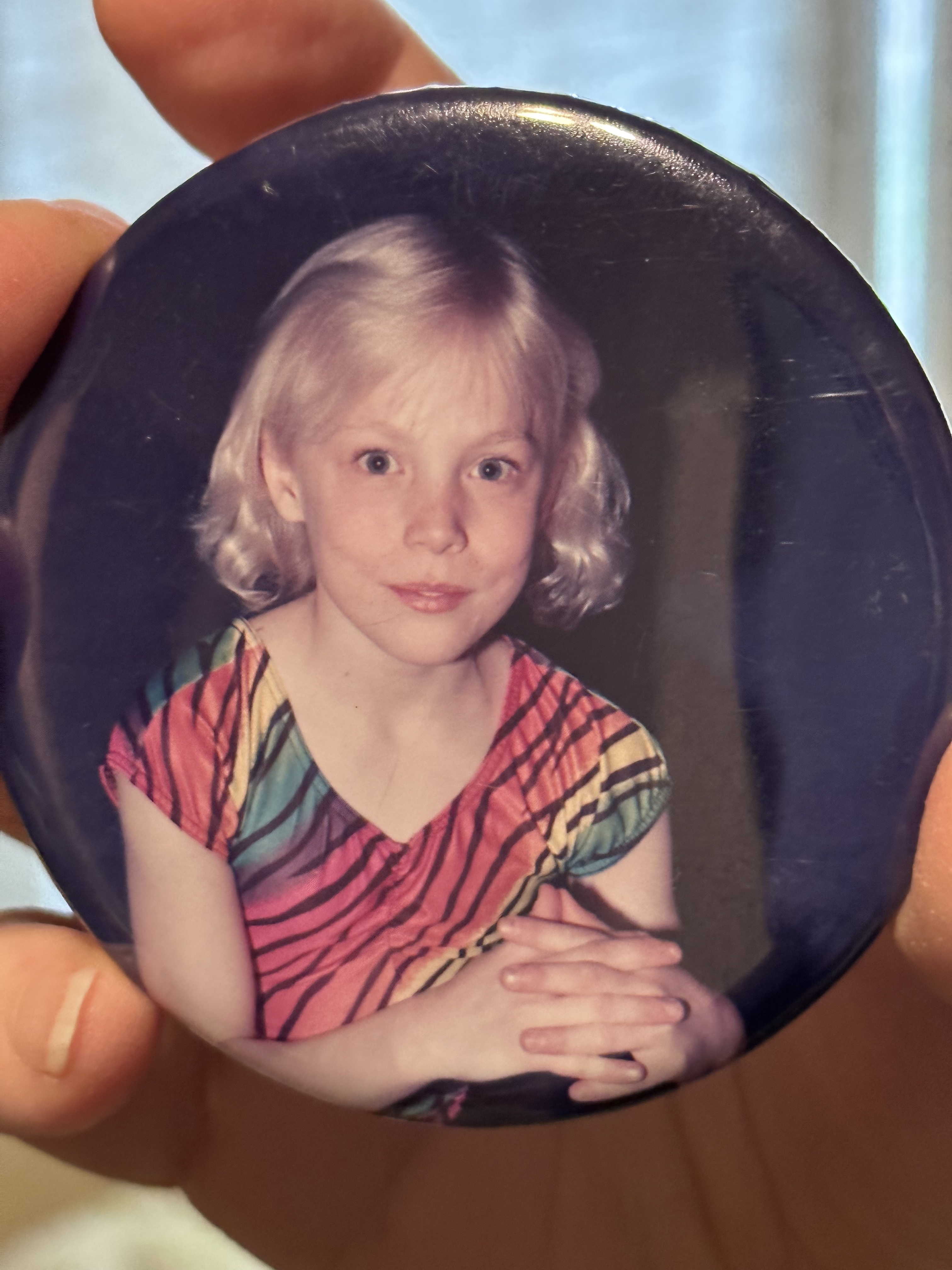 Someone holds an old button photo of a young gymnast Teo. Teo stares awkwardly into the depths of the viewers sole wearing a very Lisa Frank rainbow and tiger striped leotard.