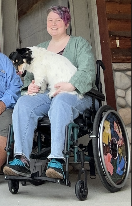 Teo sits in their wheelchair with Gracie pup on their lap. Teo is wearing a green linen top and denim jeans. Teo's wheelchair is showing wheelcovers with birds in bright colors painted on them.