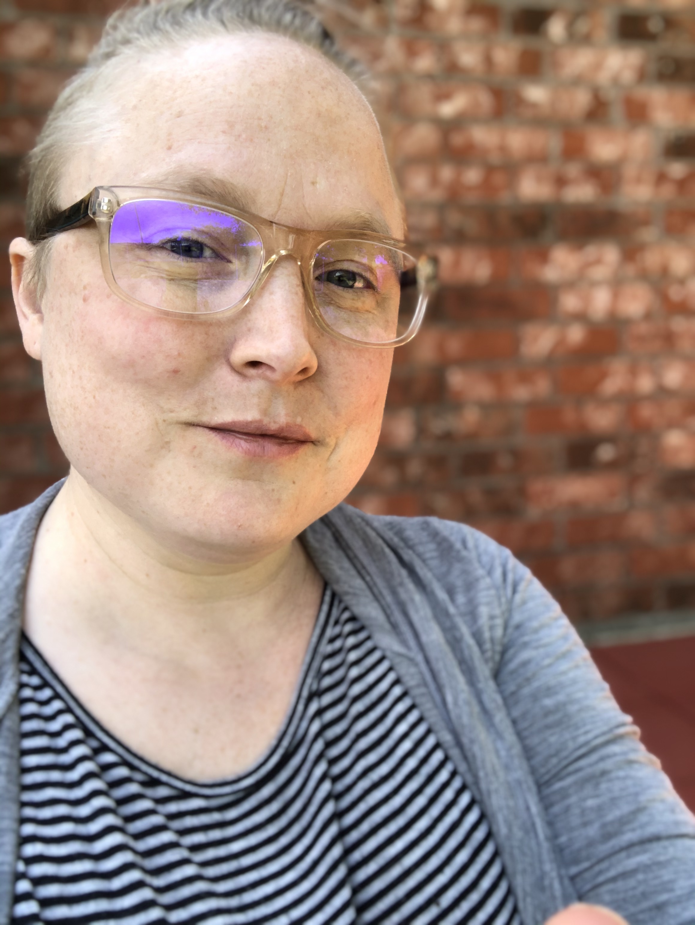 Teo with hair pulled back and clear glasses on has a small smile on, sitting in front of a brick wall. Teo wears a striped shirt and gray cardigan.
